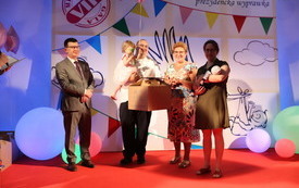 Na scenie czw&oacute;rka radosnych os&oacute;b. jedna trzyma wyprawkę dla dziecka od prezydenta. Prezydent stoi wraz z nimi z uśmiechem