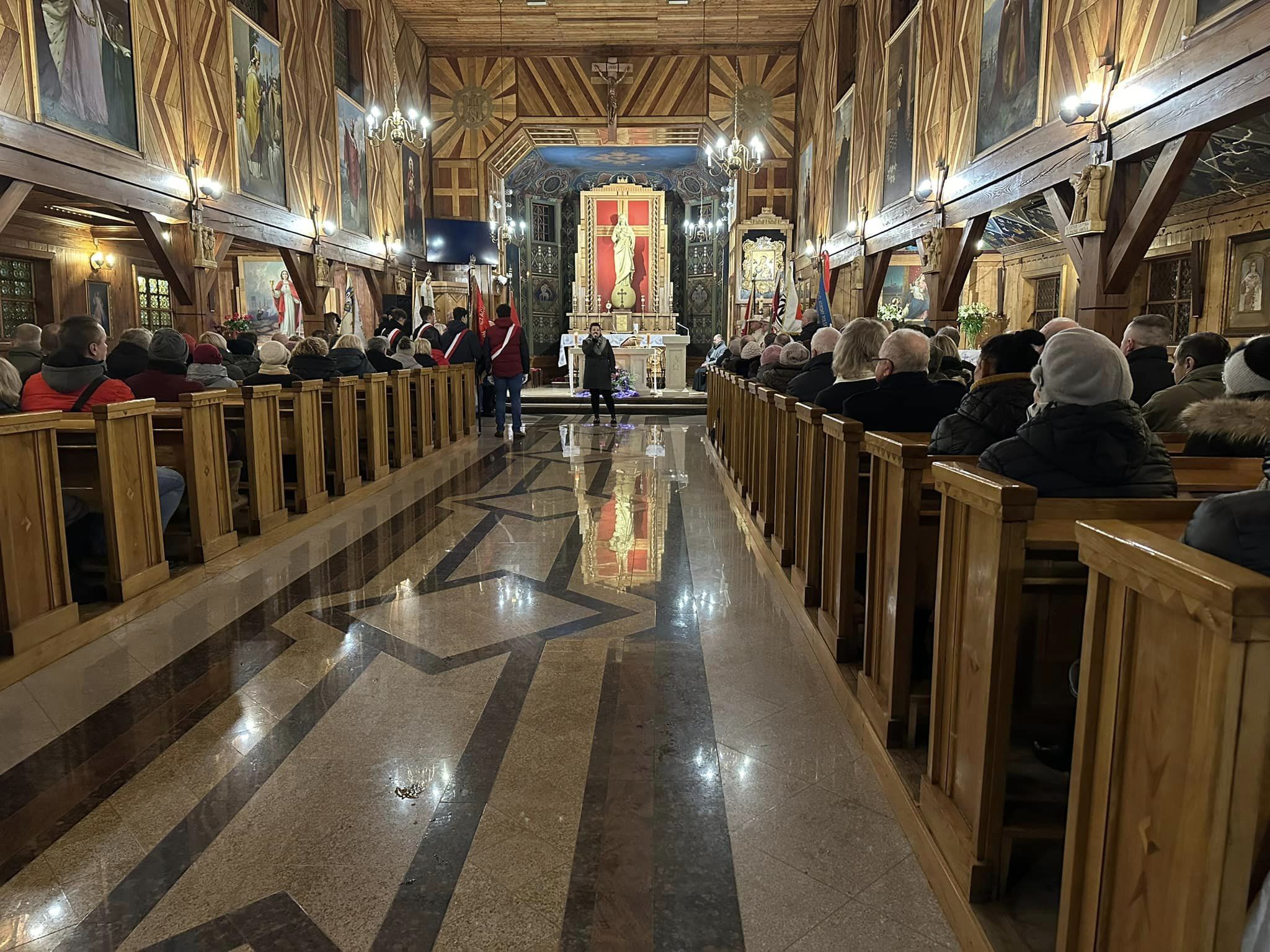 Na zdjęciu widać wnętrze kościoła, osobę stojąca na środku która śpiewa. W ławkach kościoła siedzą ludzie, przy ołtarzu ustawione są poczty sztandarowe. 