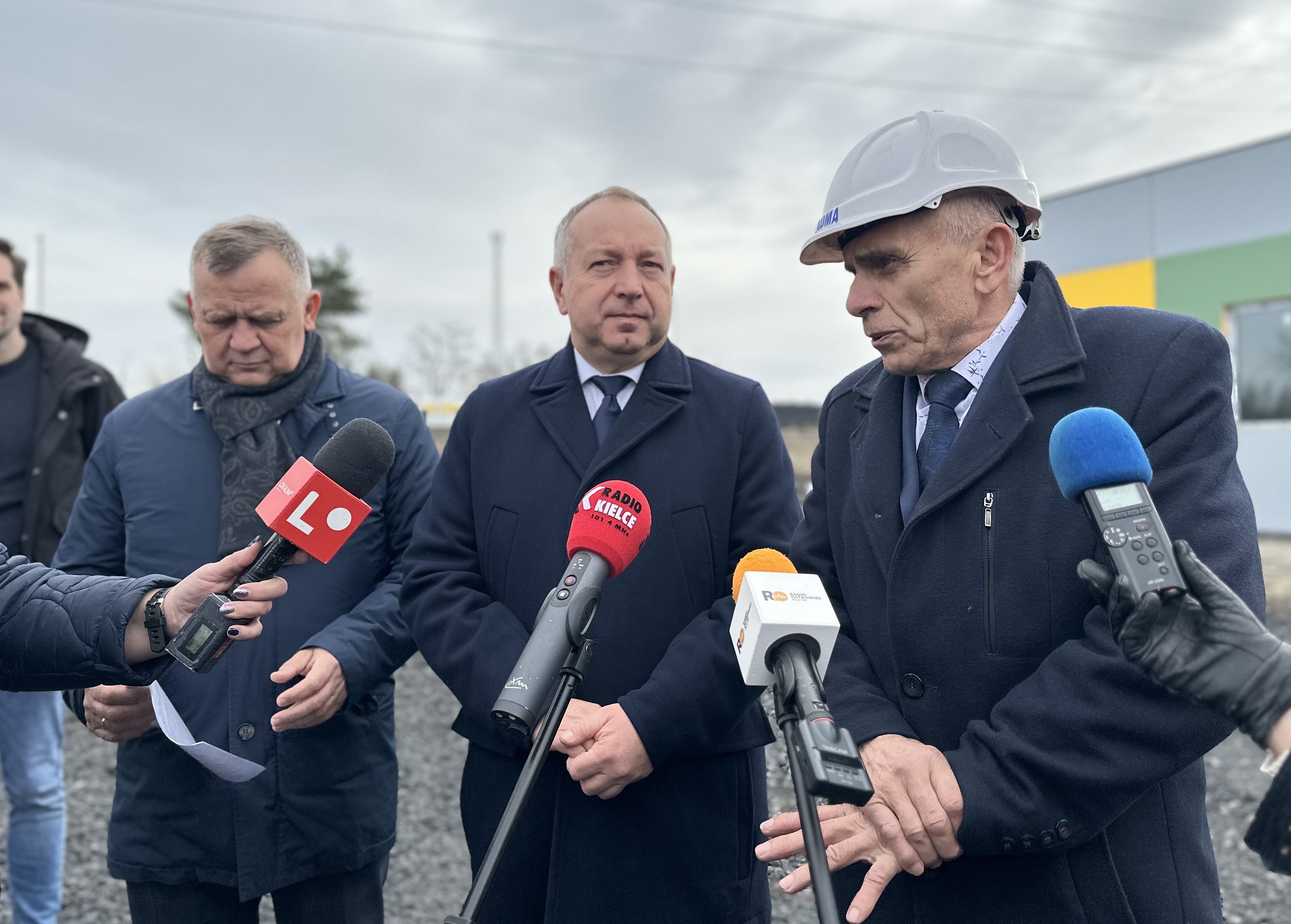 Na zdjęciu widać trzech mężczyzn w średnim wieku, znajdują się oni na placu budowy. Udzielają wywiadu mediom, więc przed nimi widać mikrofony prasowe.